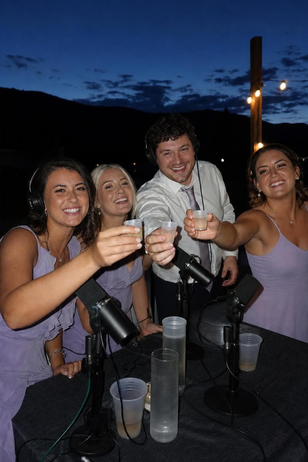 Bridesmaids and a groomsman in headphones raising a toast at the podcast table at dusk