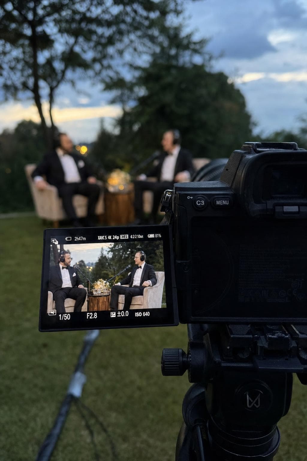 A cinema camera on a tripod framing two groomsmen in armchairs on its screen, outdoors at golden hour